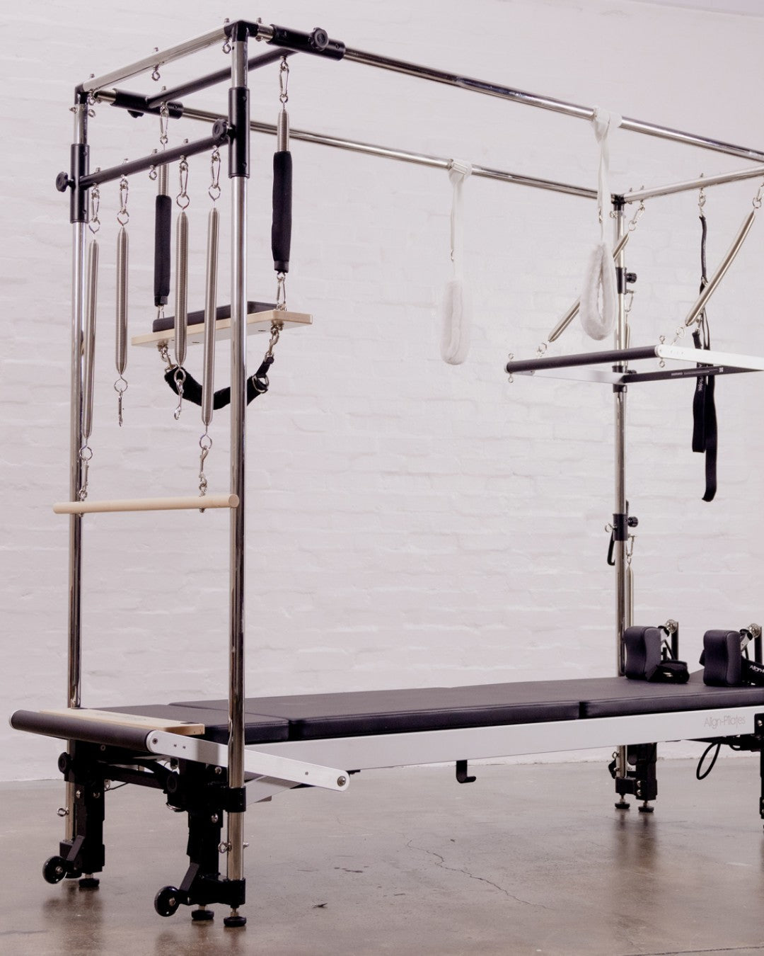 Pilates Cadillac mit Federn, Stangen und Schlaufen vor weißer Ziegelwand, davor ein Reformer