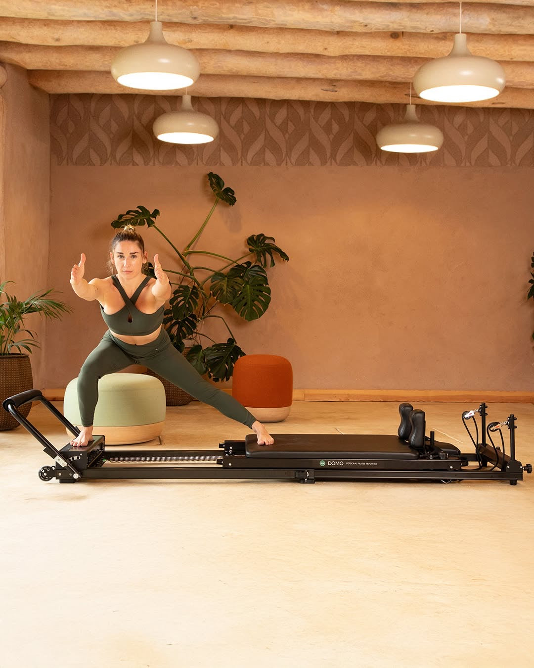Frau trainiert auf einem Pilates-Reformer in einem wohnlichen Raum mit Holzdecke und Pflanzen